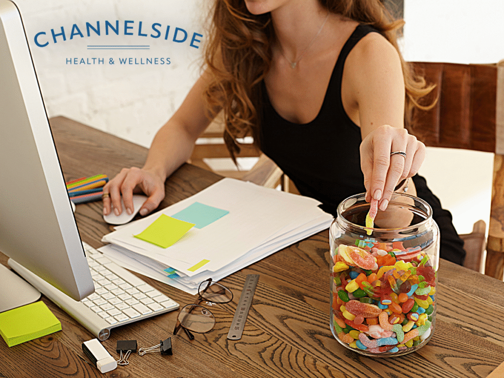 A woman working at her desk reaches into a large jar of candy, demonstrating the concept of stress eating and its impact on weight.-Stress and weight
