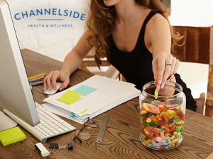 A woman working at her desk reaches into a large jar of candy, demonstrating the concept of stress eating and its impact on weight.-Stress and weight