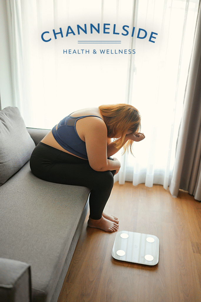 A distressed woman sits on a couch with her head in her hands and a bathroom scale on the floor in front of her, symbolizing the stress and emotional burden of weight concerns.-stress and weight